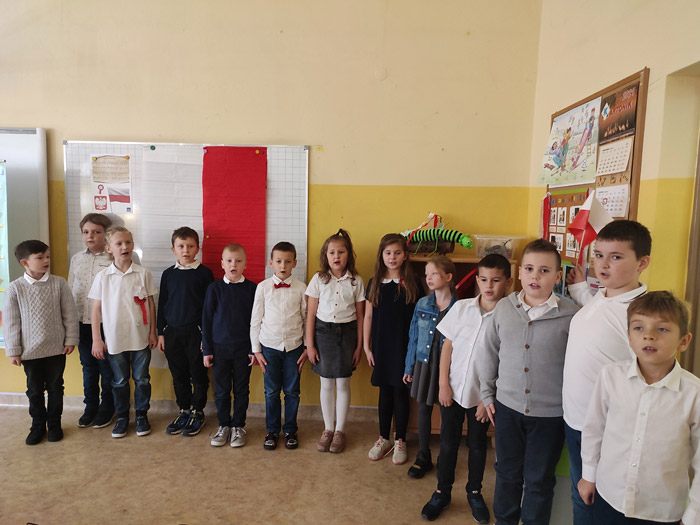 Grafika przedstawia uczniów z klasy 2a w strojach galowych podczas spiewania hymnu w klasie o godz. 11:11