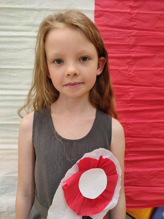 Grafika przedstawia dziewczynke z klasy 2 z kotylionem na piersie na tle biało czerwonych barw