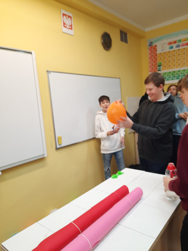Grafika przedstawia ucznia z klasy 8c przebijającego balona z wróżbą andrzejkową
