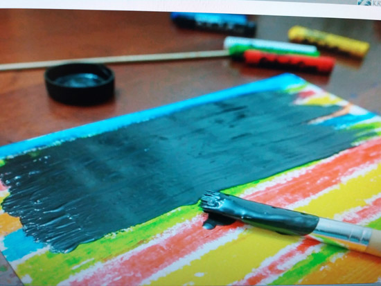 Grafika przedstawia kartke z bloku pomalowaną w paski kolorowymi pastelami, która w części pokryta jest juz czarna farbą