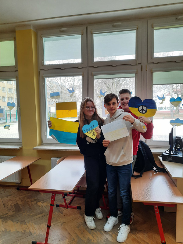 Grafika przedstawia troje uczniów z klasy 8c w sali lekcyjnej z flagami Ukrainy