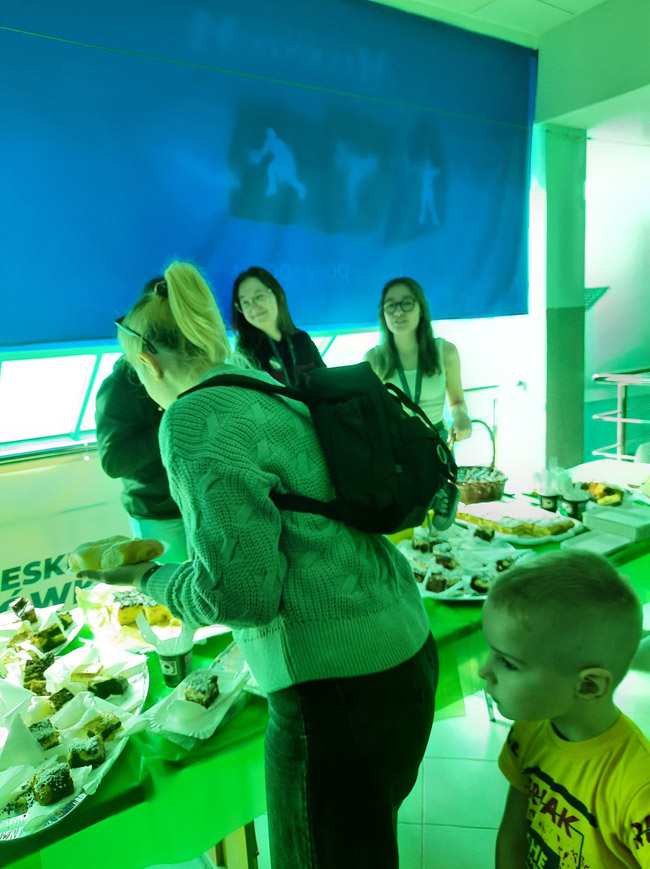 Grafika przedstawia wolontariuszy przy stole zastawionym smakołykami na Hali Milenium podczas pikniku integracyjnego