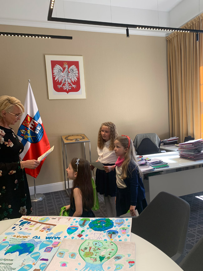 Grafika przedstawia dziewczynki nagradzane przez paniom prezydent Kołobrzegu