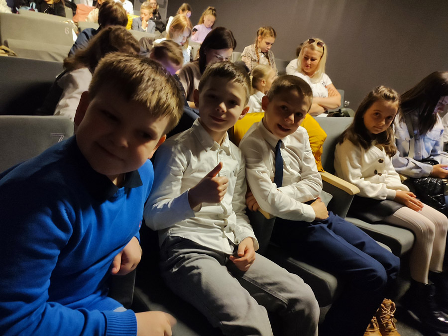 Grafika przedstawia chłopców siedzących na sali kinowej  w Regionalnym Centrum Kultury w Kołobrzegu