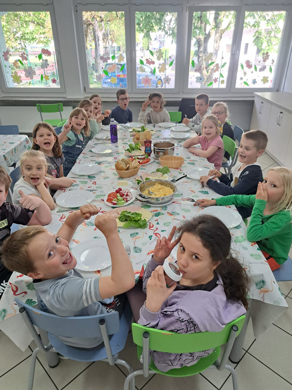 Grafika przedstawia dzieci z klasy 1c spożywające wspólnie przy jednym stolem śniadanie
