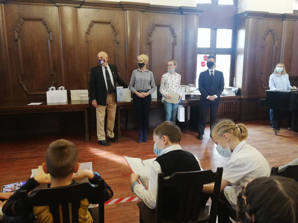Grafika przedstawia uczniów w sali koncertowej ratusza Kołobrzeskiego oraz panią prezydent miasta